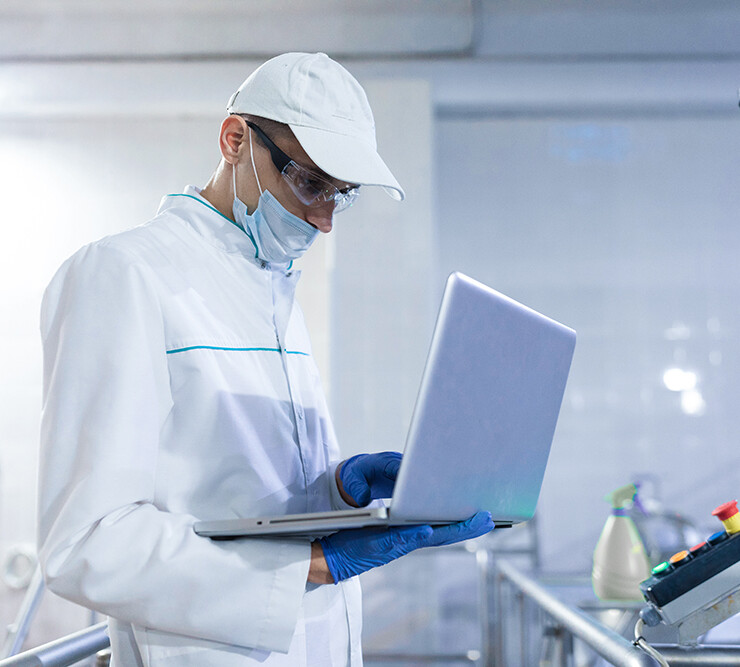 Portrait of man in a white robe and a cap standing in production department of dairy factory with laptop